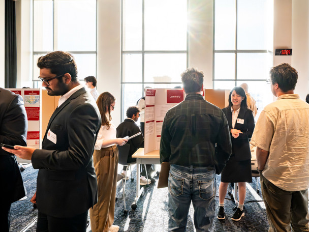 In a brightly lit exhibition hall, students stand by their posters to explain their work as visitors mill through.