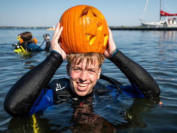 A student in scuba gear floats in the water and wears his carved pumpkin over his head like a hat.