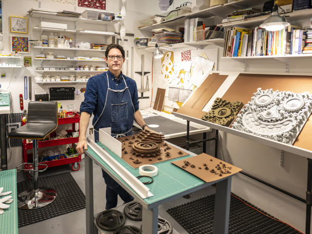 Vellliquette stands in a workroom filled with paper and art materials. He leans on a craft table that holds materials for his latest paper work.
