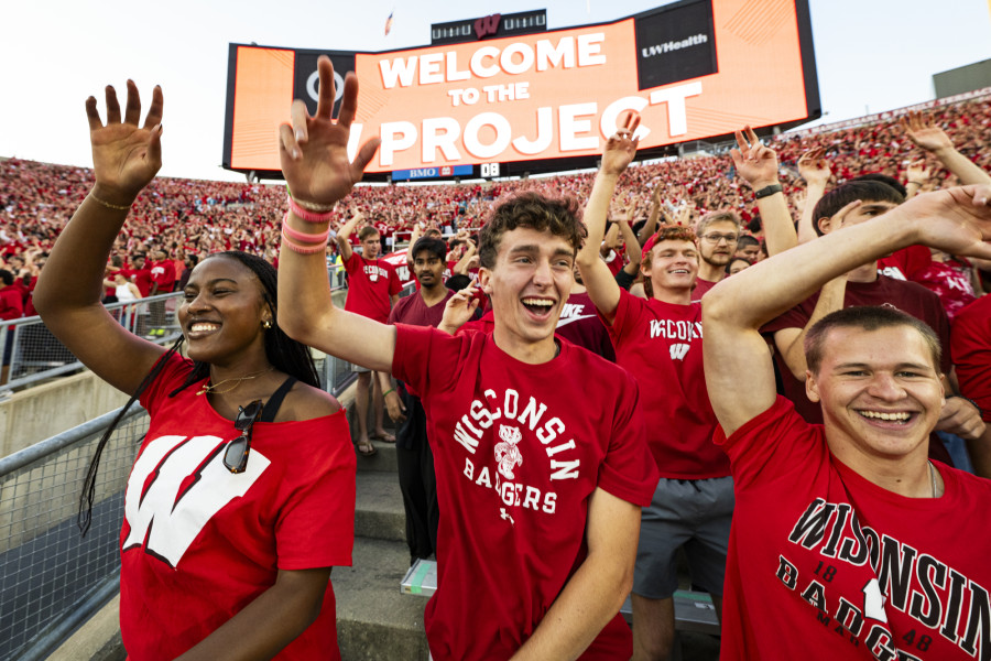 Students wearing Wisconsin Badger T shirts smile and wave as they cheer. They're in stands at a stadium.