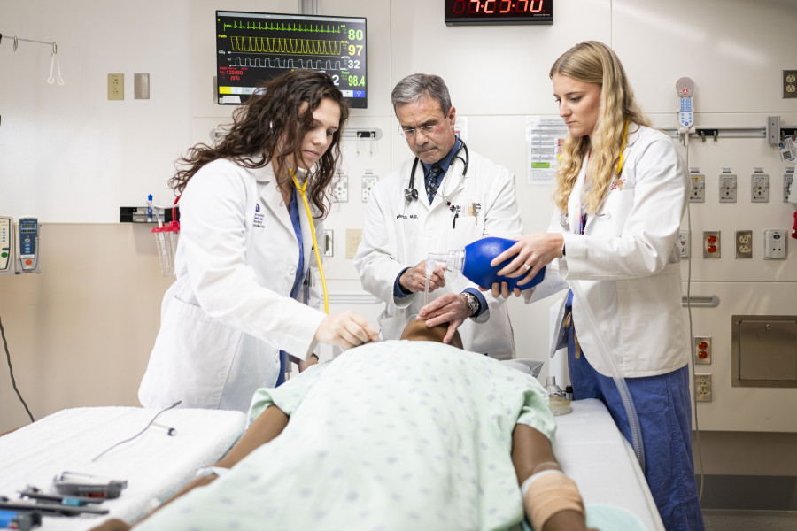 Doctor Michael Medich demonstrates procedure on medical dummy lying on a gurney to two medical students, all three wearing white lab coats