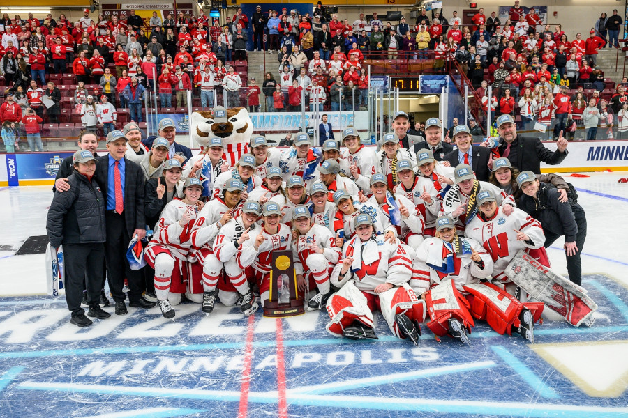The hockey team, joined by key coaches and mascot Bucky Badger, gather in a group pose on the center of the ice. The team sits behind the NCAA national championship trophy.
