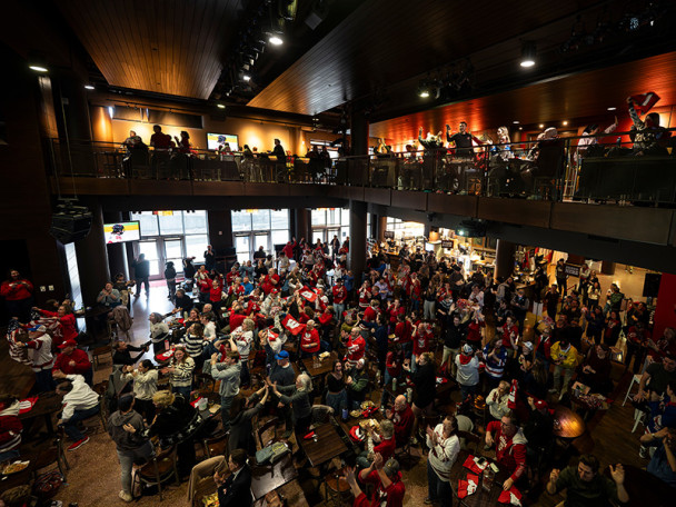 A photo taken from above shows Badger fans wearing red jerseys filling the open space of The Sett below as they watch a large screen projecting a hockey game.