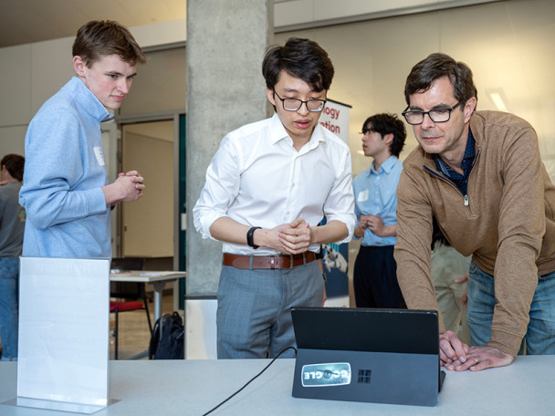 Hausman leans over a table that holds an open laptop. Wu and Cao stand next to Hausman as they explain their project concept to him.