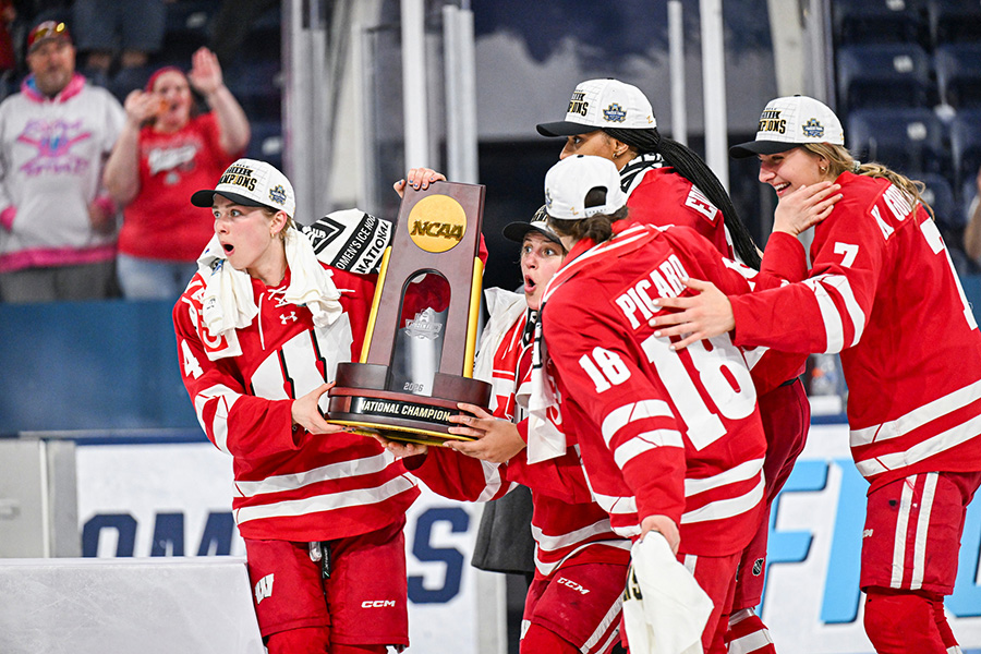 Five members of the Badger women’s hockey team hold up the NCAA championship trophy while their faces sport shocked expressions