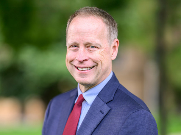 Portrait photo of John Zumbrunnen smiling. He is wearing a blue suit and red tie and standing outdoors in front of a blurred background of green trees.