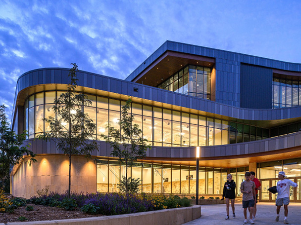 A large, modern building with many windows emanates light as dusk falls.