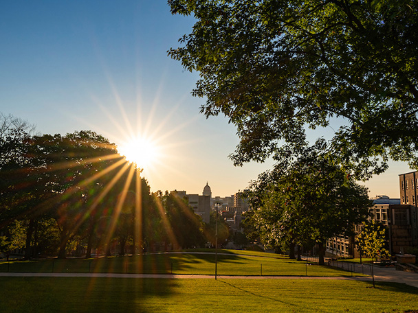 Sunrise beaming over Wisconsin State Capitol Building in distance and a treelined Bascom Hill in foreground.