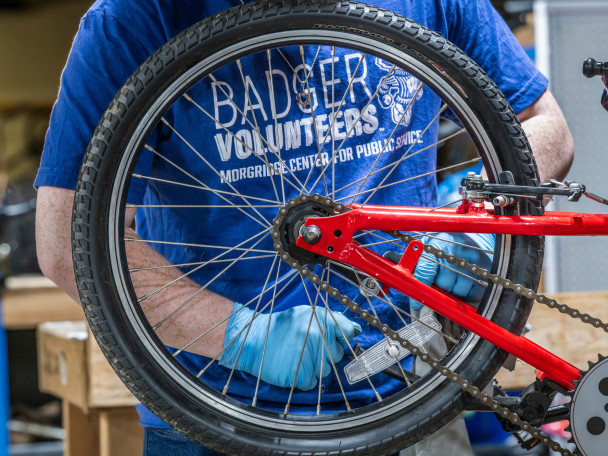 A person wearing blue gloves adjusts the rear wheel and chain of a red bicycle, with a blue shirt reading “Badger Volunteers, Morgridge Center for Public Service” visible in a workshop setting.