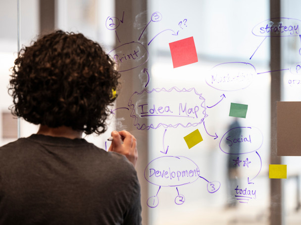 A women stands facing a see through white board. She makes notes on the board and adds colorful post it notes to the board.
