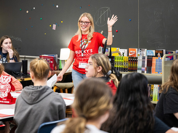Stephanie Kressin, wearing a red 'Bucky's Classroom' t-shirt, stands up in front of a class and talks and waves.