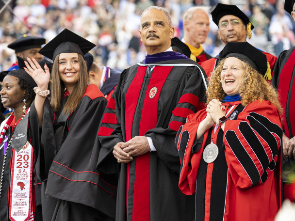 Three people in academic regalia stand and wave to the crowd in a stadium.
