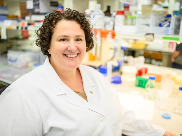Portrait of Kirstan Gimse in a white lab coat in front of a lab bench filled with colorful equipment.