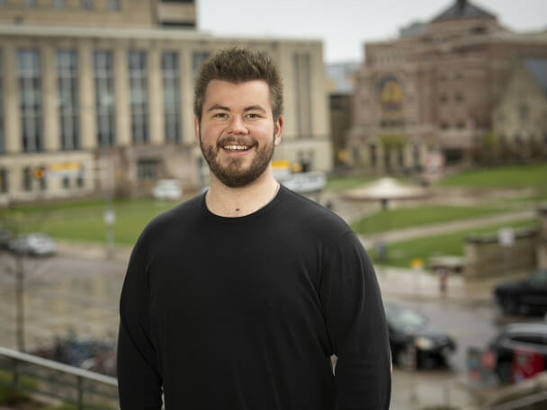 A portrait photo of Liam McLean standing outside with a green lawn and campus buildings out of focus behind him.