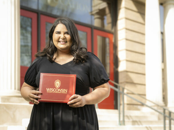 Yasmeena Ougayour stands outside the School of Education, a sandstone building with white columns and red doors. She is smiling to the camera and holding a red diploma folio with the University of Wisconsin–Madison seal and the words "Wisconsin University of Wisconsin–Madison."