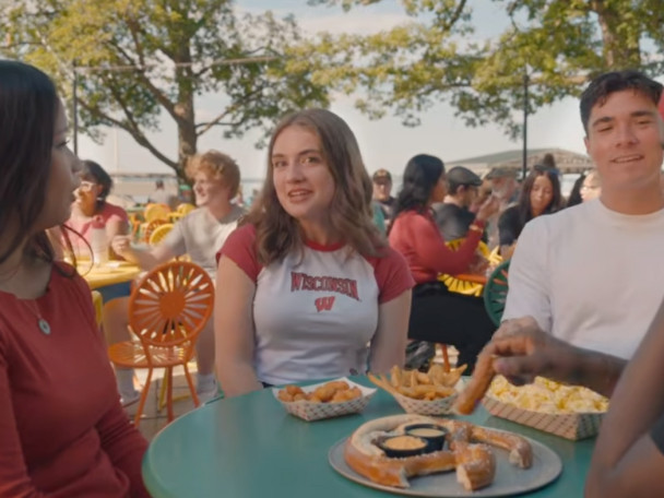 A group of young adults sit around a green outdoor table filled with snacks at a lakeside terrace while a woman wearing a Wisconsin t-shirt smiles at the camera.