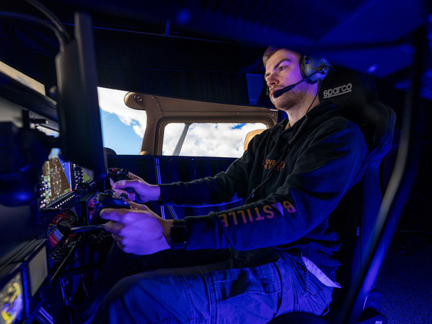 Person seated in a flight simulator cockpit, wearing headphones and using the controls, with screens showing flight instruments and a cloud-filled sky through simulated aircraft windows.