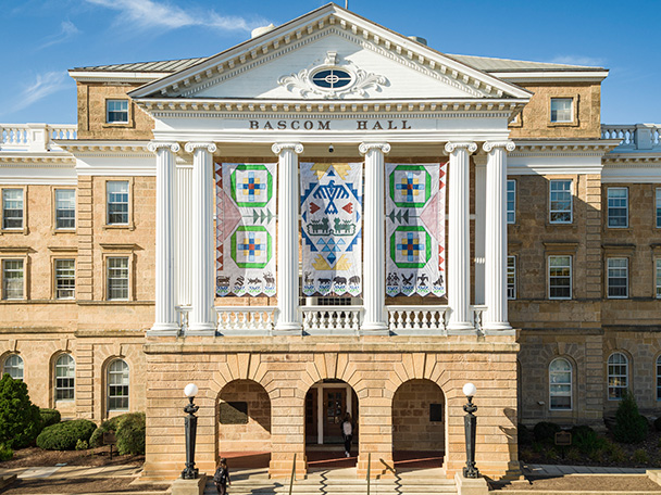 A view of the front facade of Bascom Hall taken from a drone shows three banners depicting Ho-Chunk artwork hanging between four central white columns.