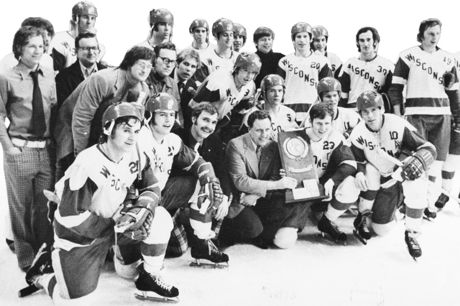 Black-and-white photo of a hockey team in Wisconsin uniforms gathered on the ice with coaches and staff, posing together while one person holds a championship trophy at the center.