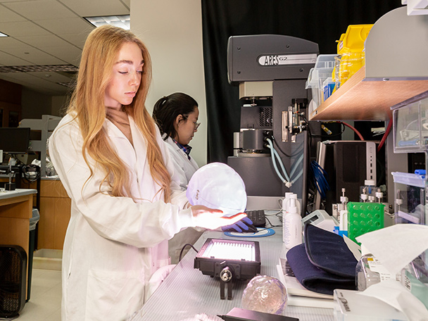 A woman in a white lab coat holds a clear 3D printed skull used for research.