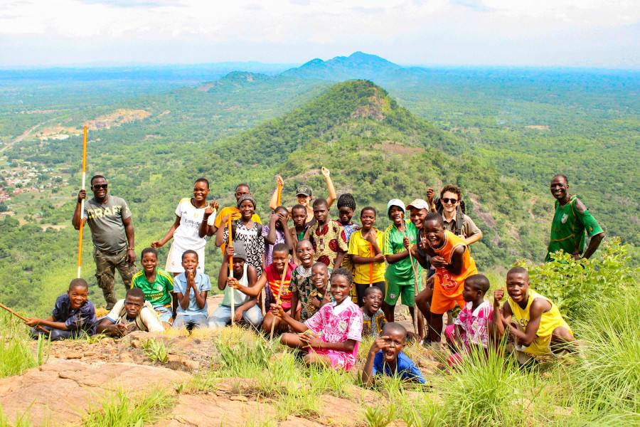 A large group of smiling children and adults pose together on a rocky hill summit overlooking an expanse of lush green countryside.