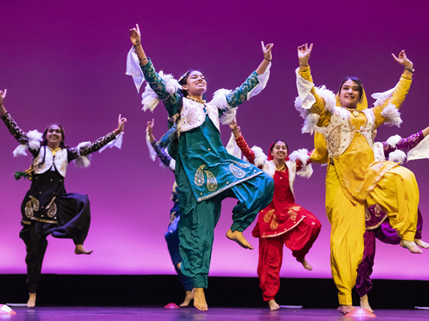 People in colorful, traditional costumes of Northern India dance on stage, a purple background behind them.