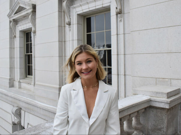 Meg Cirii stands in front of a white stone building wearing a white blazer.
