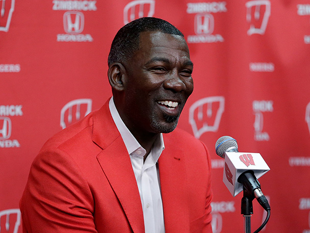Michael Finley speaking into a microphone, wearing a red blazer in front of a Wisconsin background.