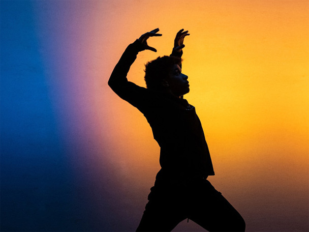 A dancer in silhouette raises his hands and arches his back in a full-body wave motion. He is backlit with blue and yellow light.