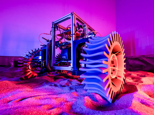 A rover with oversized, uniquely designed wheels sits on sandy terrain under vivid purple and orange lighting.