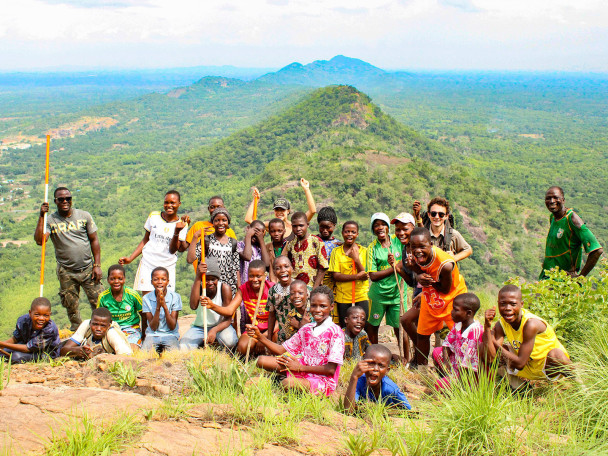 A large group of smiling children and adults pose together on a rocky hill summit overlooking an expanse of lush green countryside.