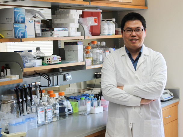 Quanyin Hu, wearing a white lab coat, stands with arms crossed in a laboratory, smiling at the camera.