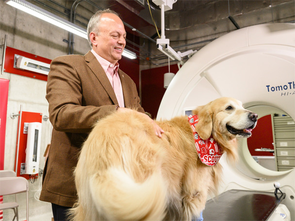 Scout, a golden retriever stands on an exam table and a medical imaging device. He is wearing a red and white bandana printed with Bucky Badger, the Wisconsin W and his name. His owner, WeatherTech Founder and CEO David MacNeil, stands at his side smiling.