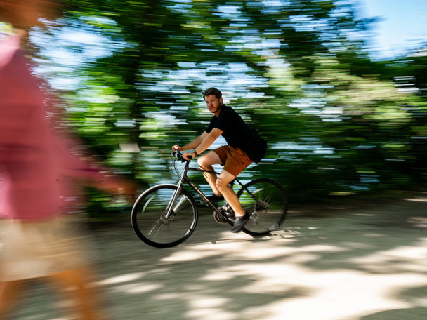 A cyclist, in focus, rides on a gravel path, a blurry, tree-lined background behind him.