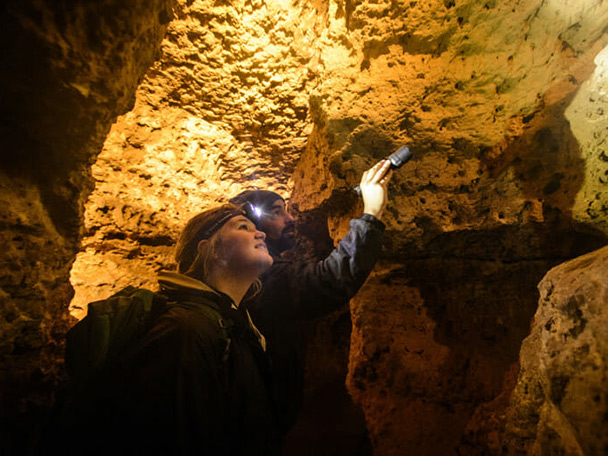 A man and woman stand in a well-lit cavern. The man holds a small flashlight and points to something above their heads.