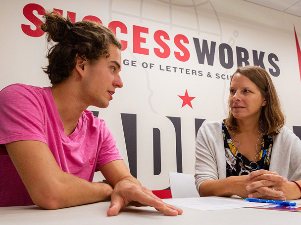 L&S student Colton Mansavage discusses his future goals with SuccessWorks’ interim director Angie White in front of a colorful wall with the Success Works logo.