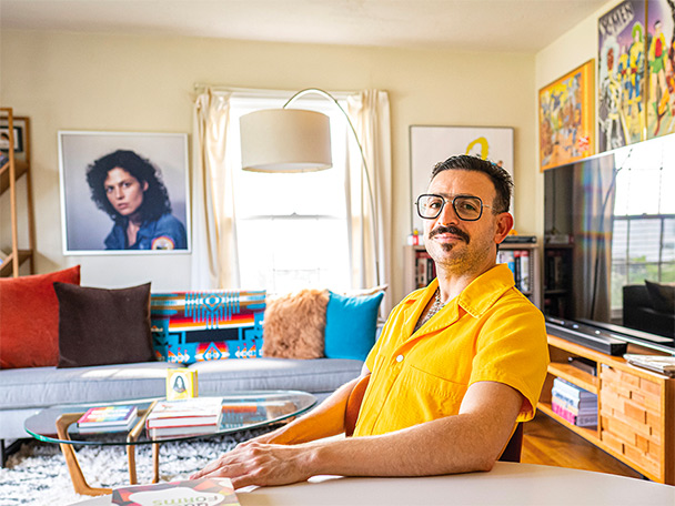 Ramzi Fawaz wearing a bright yellow jumpsuit, in his living room; the walls covered with colorful artwork.