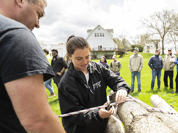 A man and a woman stand by a log and work on tying a rope to it.
