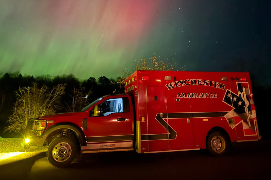 A red ambulance with the words "Winchester Ambulance" on the side of the road at night with Northern Lights in the background