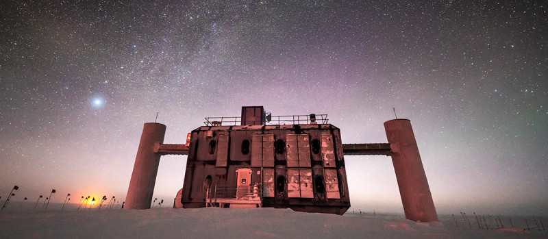 Front view of the IceCube Lab at twilight, with a starry sky showing a glimpse of the Milky Way overhead and sunlight lingering on the horizon.