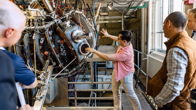 One person stands in front of a large metal machine and explains how the machine works to several people who are listening nearby.
