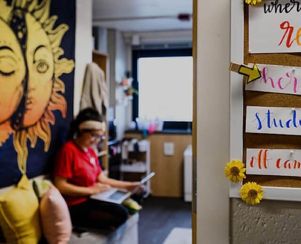 Inside a residence hall. A bulletin board is on the wall, with a student working on a laptop in the background.