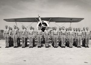 Thirty-some Members of the Flying Badgers dressed in uniform standing in front of a biplane.