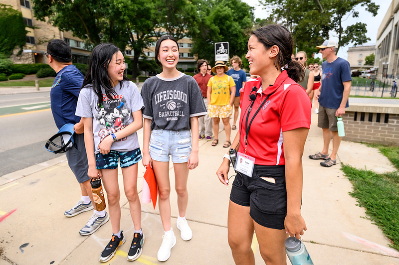 A girl in a red shirt with a lanyard smiles and talks to two girls on the sidewalk.